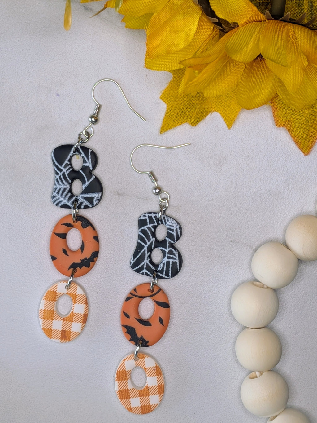 A pair of drop earrings with three stacked circular and patterned elements, featuring a black and white spiderweb design, orange with black spiderweb print, and orange and white gingham spelling out the word "Boo." The earrings are displayed on a textured white surface with yellow flowers and wooden beads in the background.
