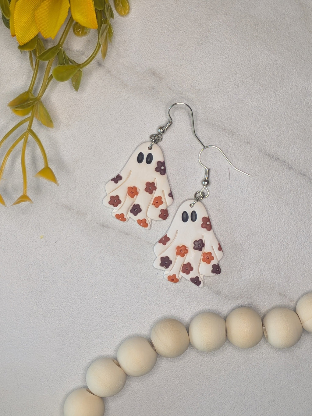 A pair of ghost-shaped dangle earrings with a white body, black eyes, and a floral pattern in orange, red-brown, and burgundy, displayed on a white marble surface with a sunflower and wooden beads as props.