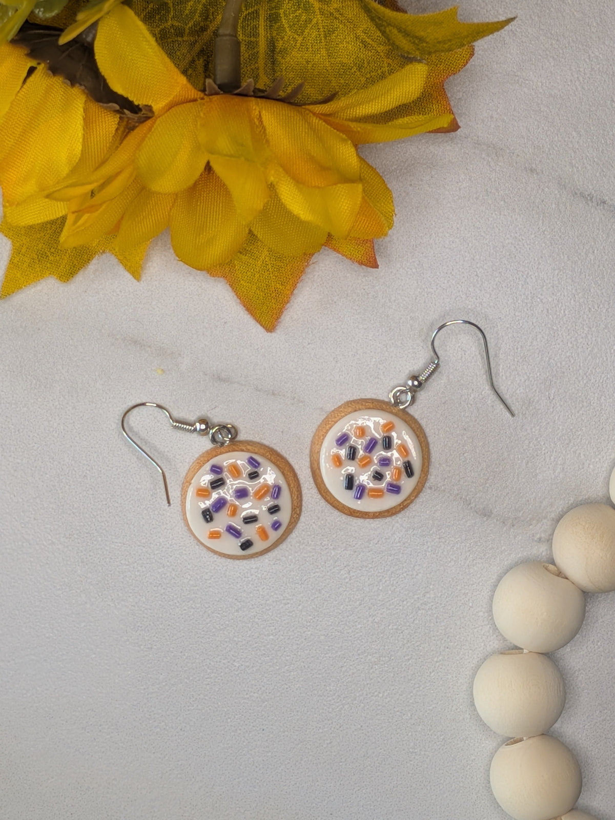 A pair of round drop earrings with silver fishhook wires shaped like sugar cookies with thick white frosting and colorful sprinkles in orange, purple, and black on a white background. The earrings are styled on a white marble surface next to yellow autumn leaves and a wooden bead strand.