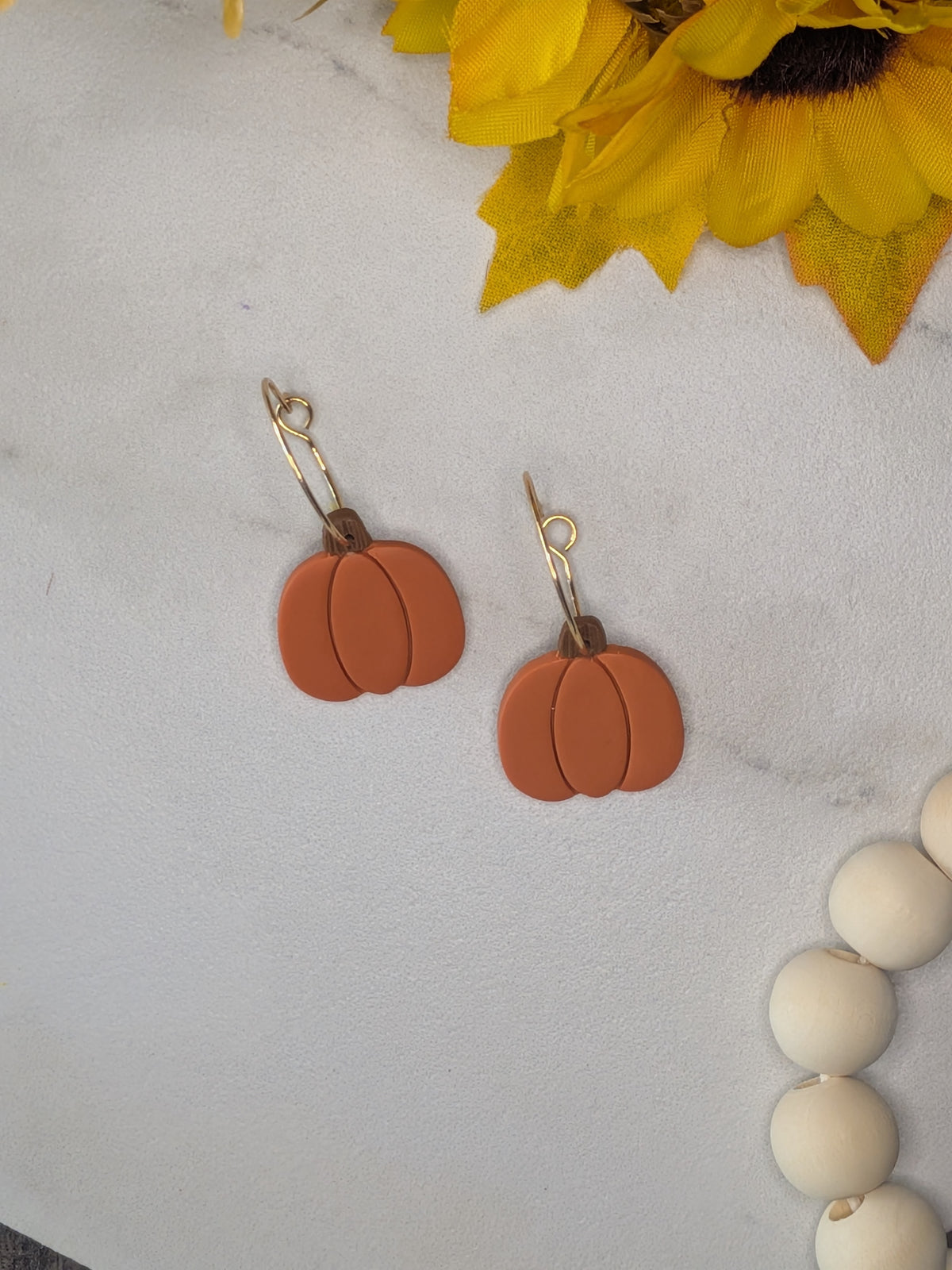 Pumpkin-shaped drop earrings with a matte terracotta finish and gold plated hoop wires, displayed on a light gray marble surface with decorative sunflower and wooden beads.
