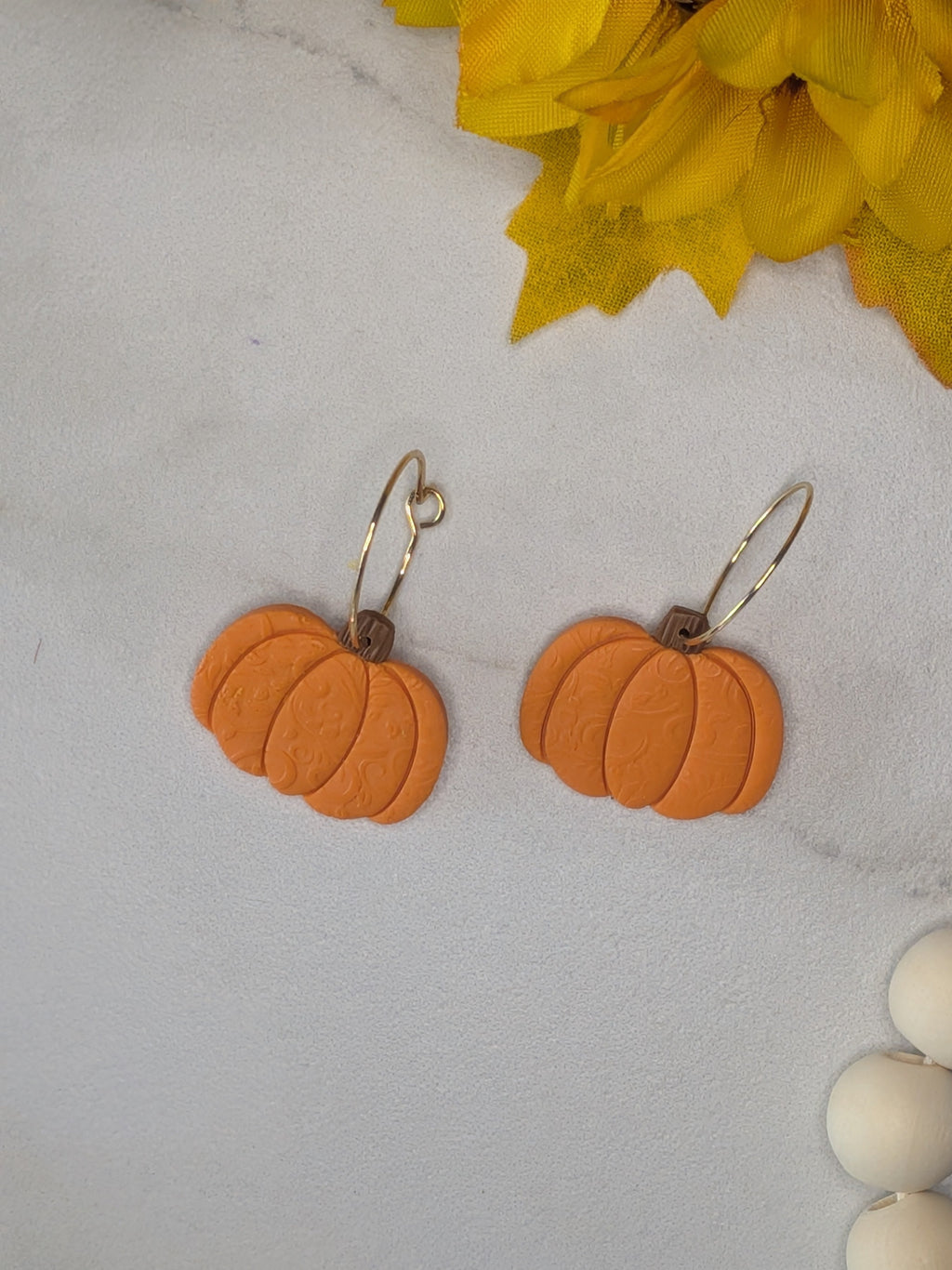 Orange pumpkin-shaped dangle earrings with a subtle swirl texture on gold plated wire hoops, displayed on a white marble surface, with yellow flowers and white beads as props.