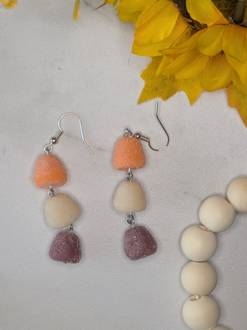 A pair of dangling earrings with three stacked gumdrop shaped beads in orange, cream, and dusty mauve on silver fishhook wires, displayed on a textured white surface with yellow flowers and a beaded necklace prop.