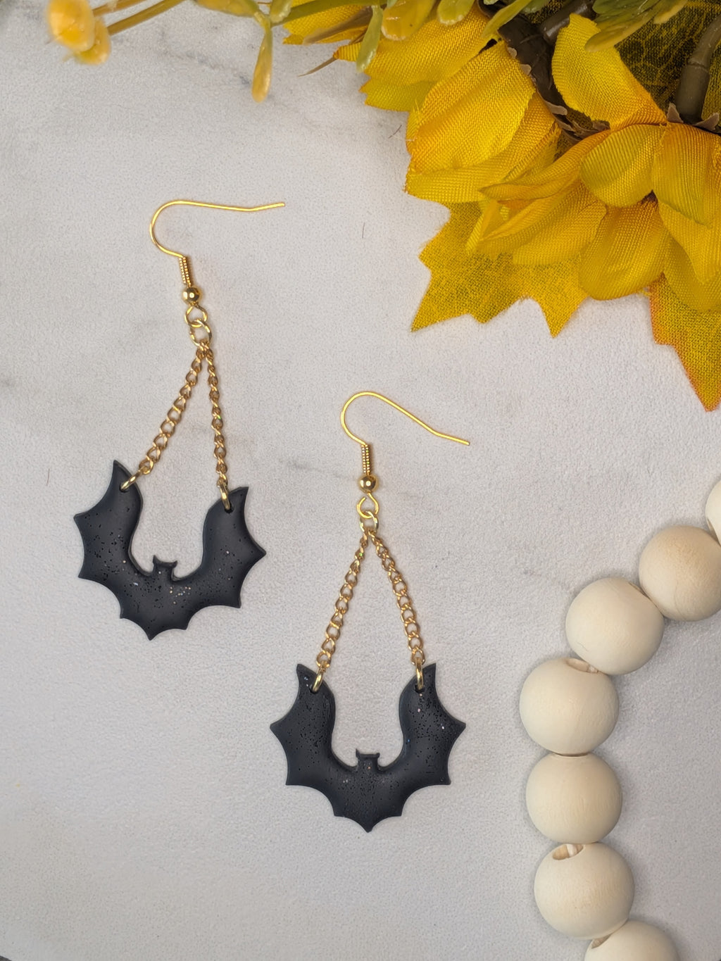 A pair of black bat-shaped drop earrings with black glitter on gold-tone chains and fishhook wires, displayed on a light textured surface with yellow flowers and white wooden beads as props.