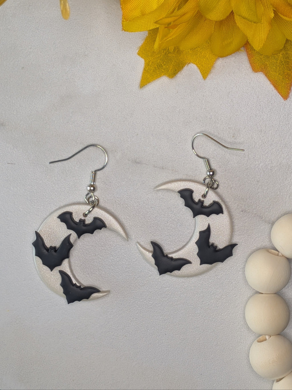 A pair of crescent moon dangle earrings with black bat silhouettes on a white background, displayed on a light marble surface with yellow flowers and a wooden bead strand nearby.