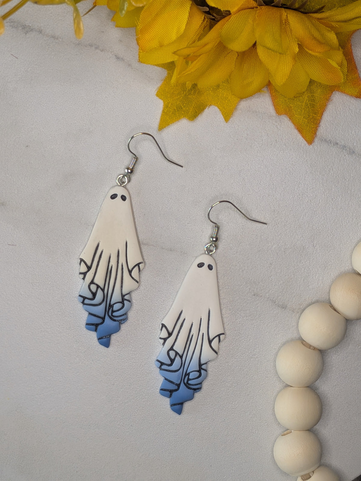 A pair of dangling ghost-shaped earrings with a white top and blue gradient bottom, displayed on a light marble-like surface with yellow leaves and white beads as props.
