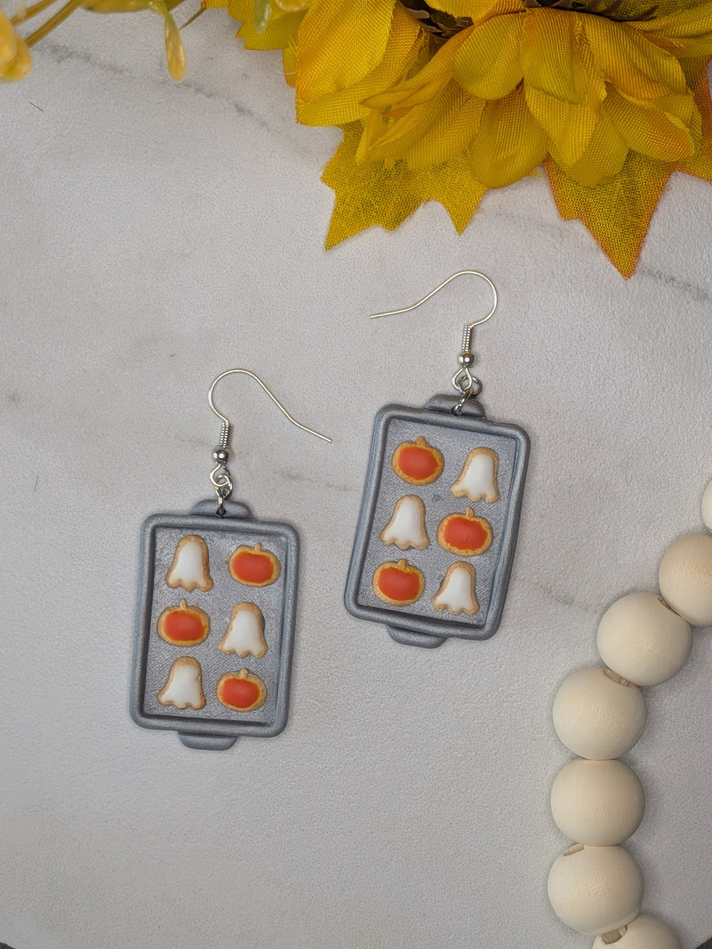 A pair of rectangular silver-tone earrings shaped like miniature baking trays, featuring orange pumpkin and white ghost cookie motifs, displayed on a light marble surface with yellow flowers and a wooden bead garland.