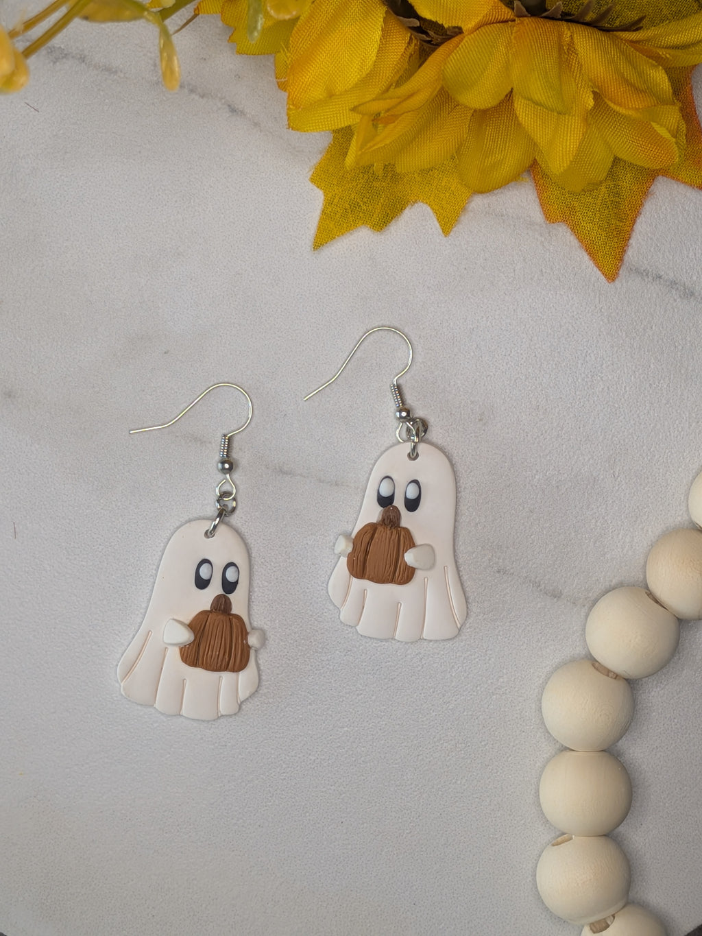 A pair of white ghost dangle earrings with brownish-orange pumpkins and silver hooks, displayed on a white marble surface with yellow flowers and a wooden bead garland.