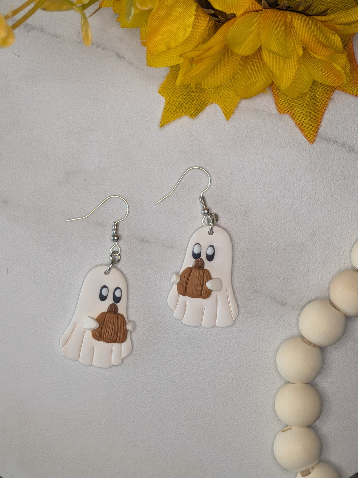 A pair of white ghost dangle earrings with brownish-orange pumpkins and silver hooks, displayed on a white marble surface with yellow flowers and a wooden bead garland.
