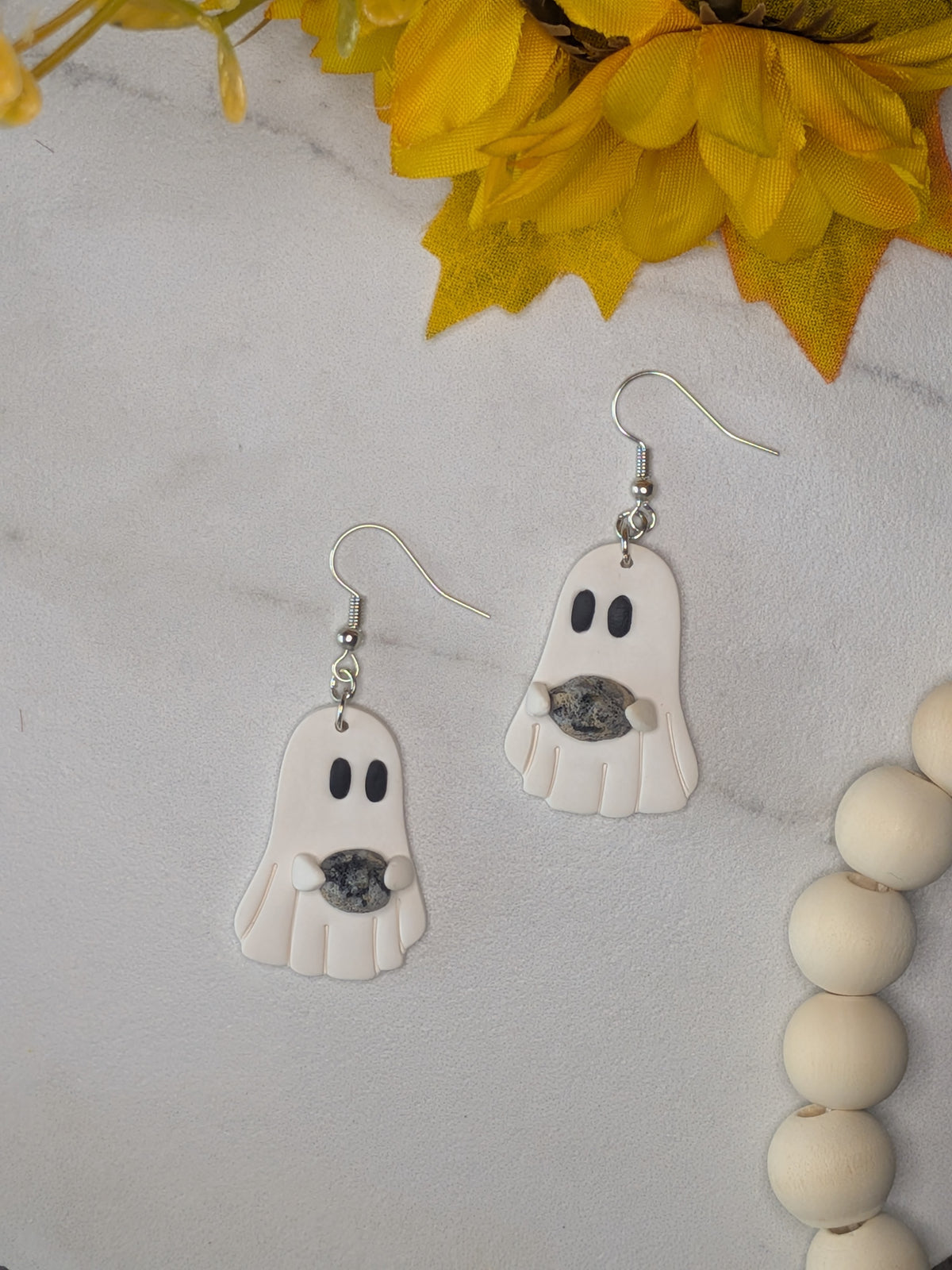 A pair of white ghost-shaped dangle earrings with black eyes, holding a black and grey rock, hanging from silver fishhook wires on a light white marble surface with yellow flowers and a wooden bead strand as props.