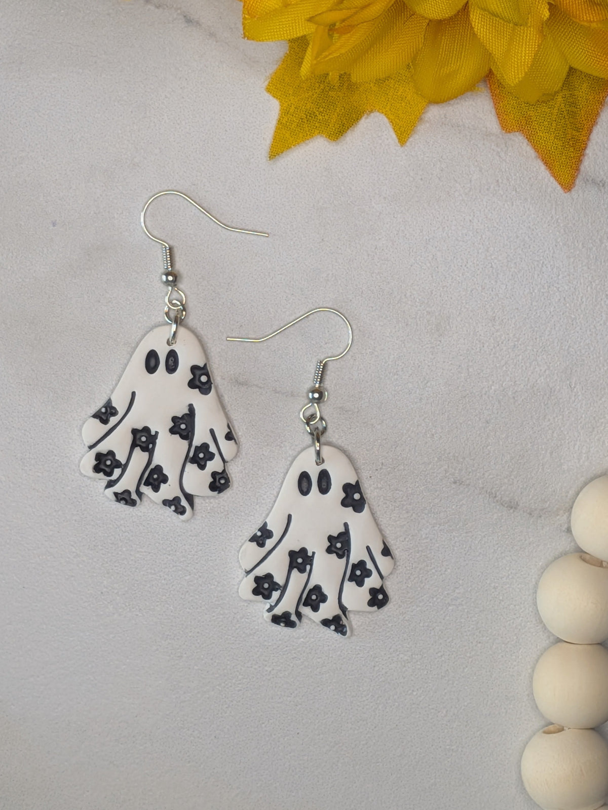 A pair of white ghost-shaped dangle earrings with black floral patterns and silver fishhooks, displayed on a light marble surface with yellow flowers and wooden beads as props.