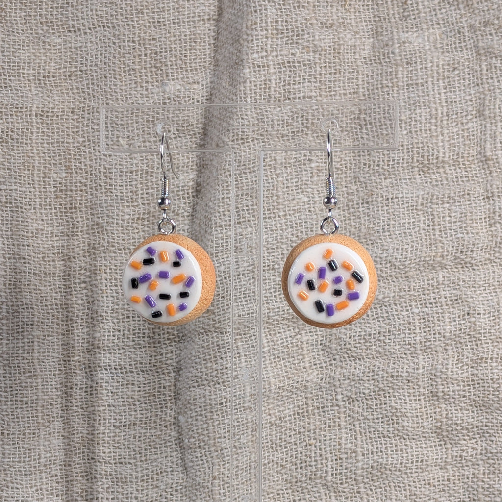 A pair of round drop earrings with silver fishhook wires shaped like sugar cookies with thick frosting and colorful sprinkles in orange, purple, and black on a natural linen background.