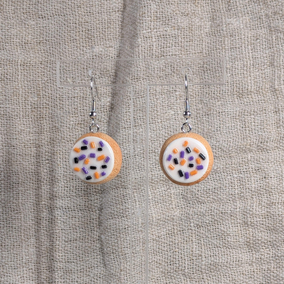 A pair of round drop earrings with silver fishhook wires shaped like sugar cookies with thick frosting and colorful sprinkles in orange, purple, and black on a natural linen background.