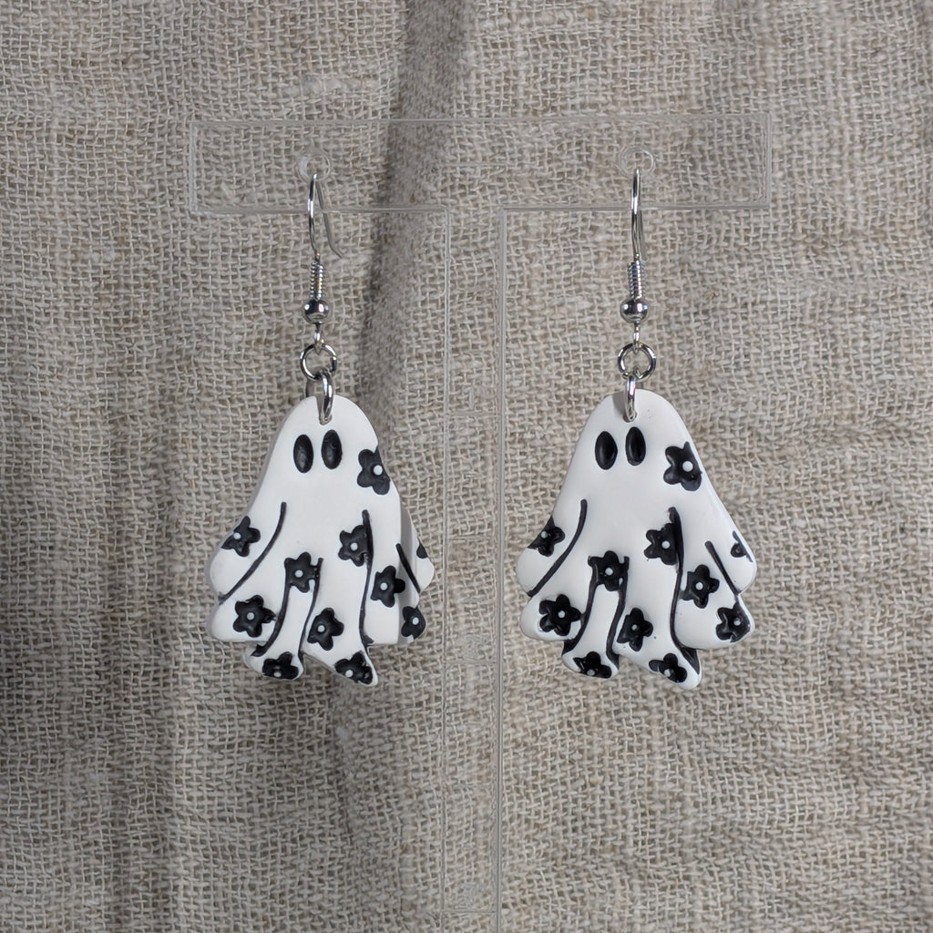 A pair of white ghost-shaped dangle earrings with black floral patterns, hanging against a textured beige burlap backdrop.