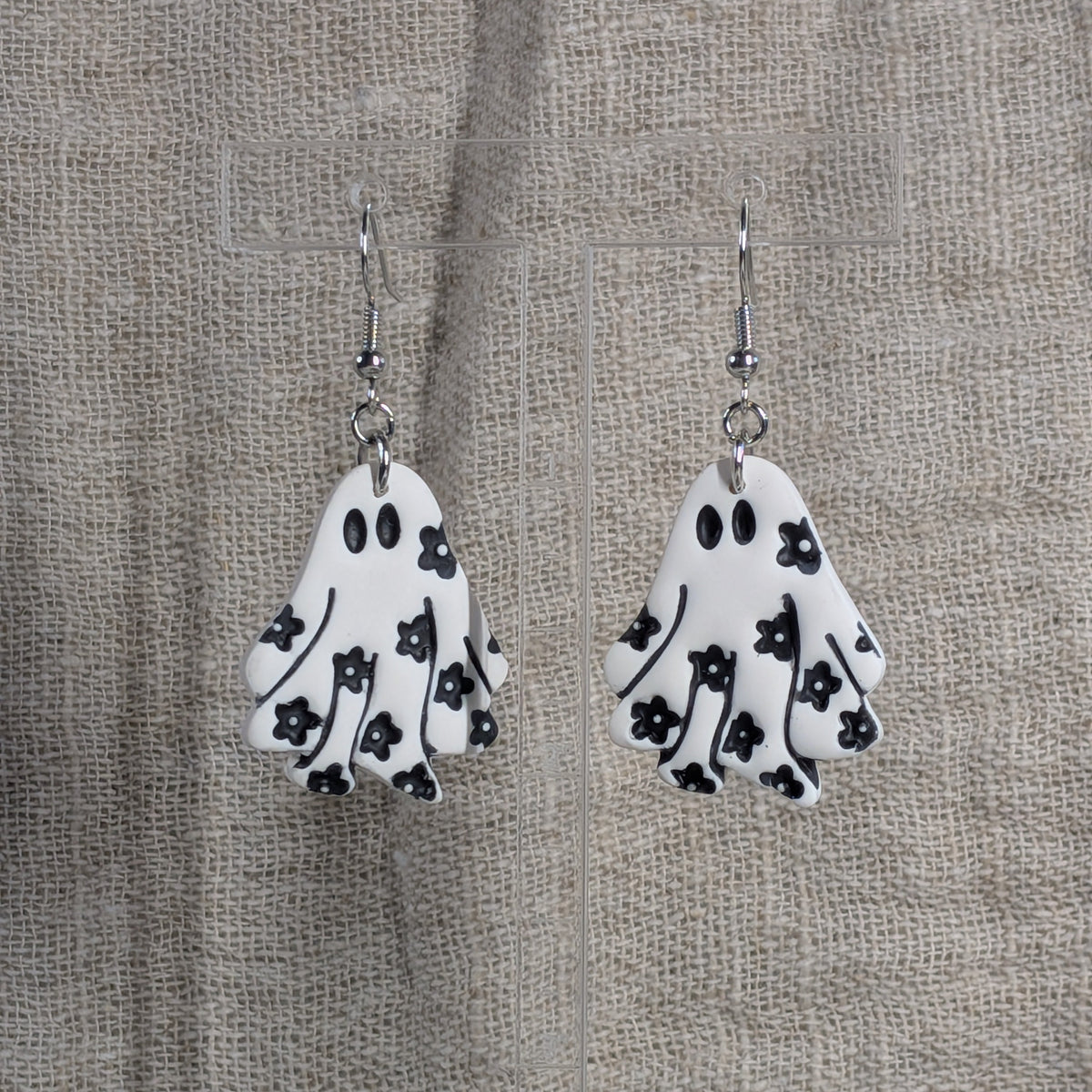 A pair of white ghost-shaped dangle earrings with black floral patterns, hanging against a textured beige burlap backdrop.