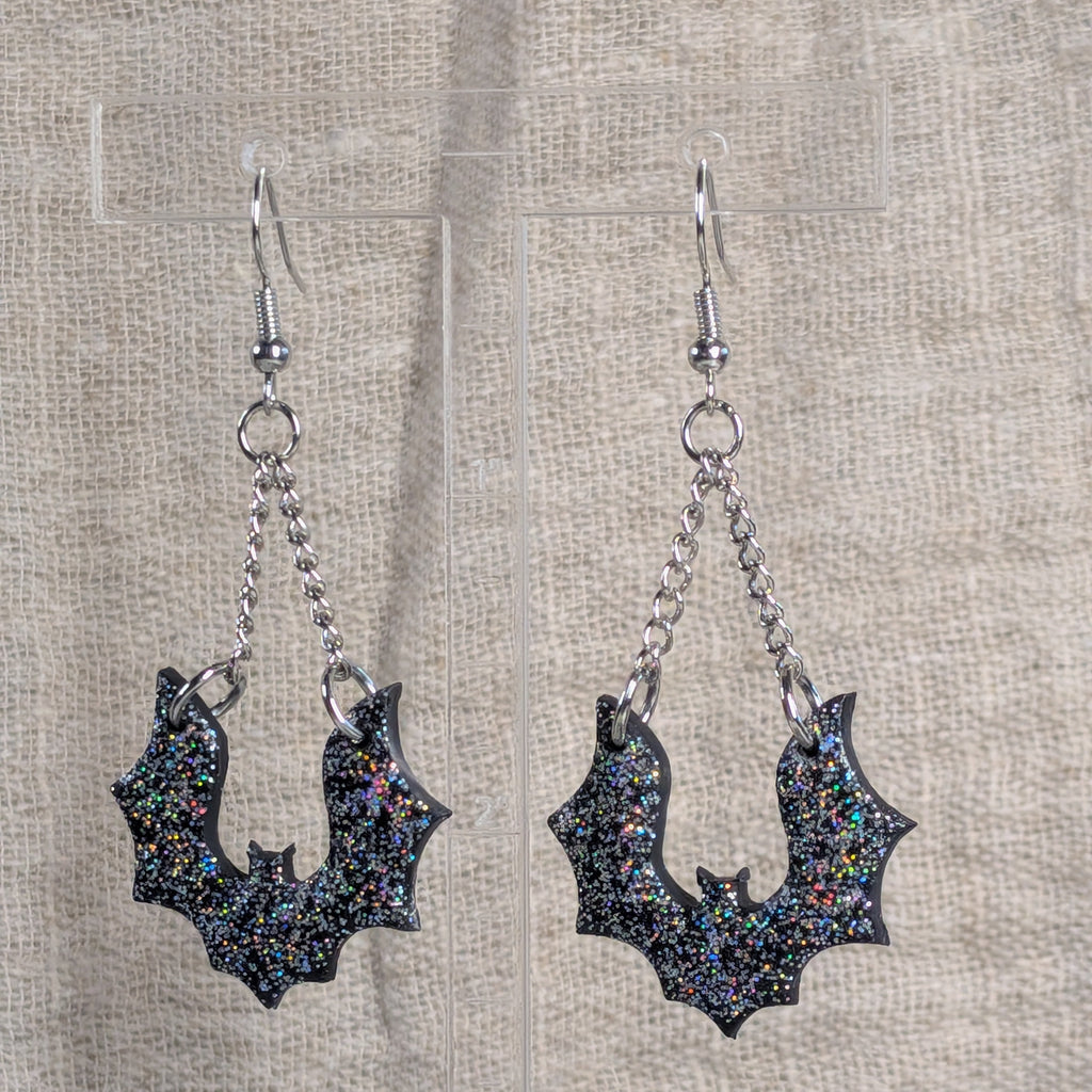 A pair of silver-toned dangle earrings with bat-shaped charms covered in black glitter and iridescent multicolor flecks, displayed on a clear stand against a beige linen background.