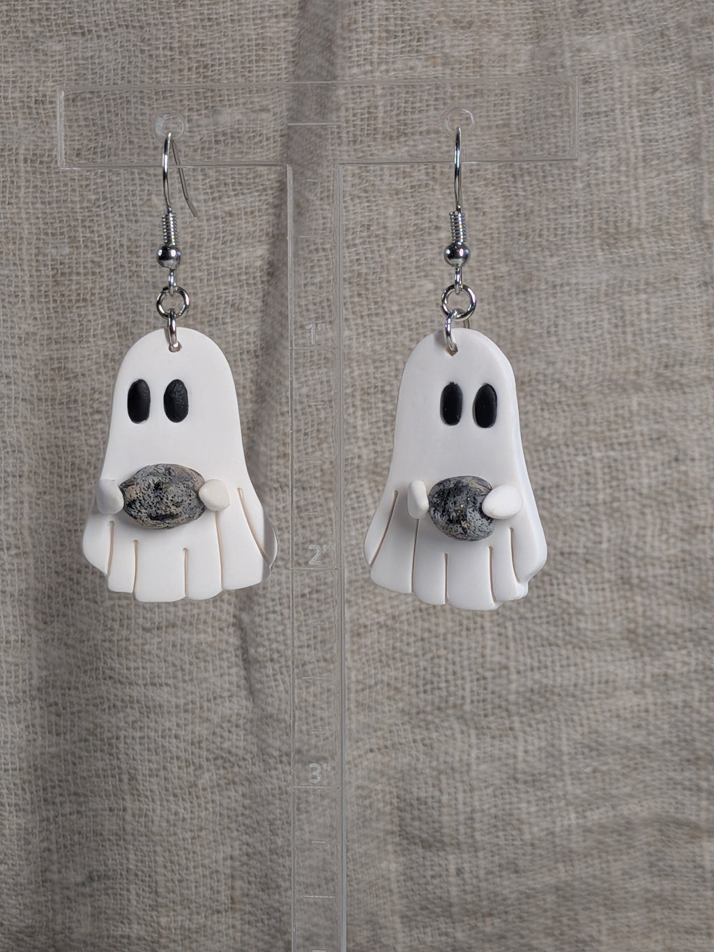 A pair of white ghost-shaped dangle earrings with black eyes, holding a small grey and black stone, hanging on silver fishhook wires against a textured beige fabric background.