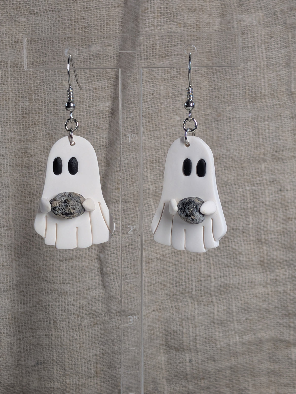 A pair of white ghost-shaped dangle earrings with black eyes, holding a small grey and black stone, hanging on silver fishhook wires against a textured beige fabric background.