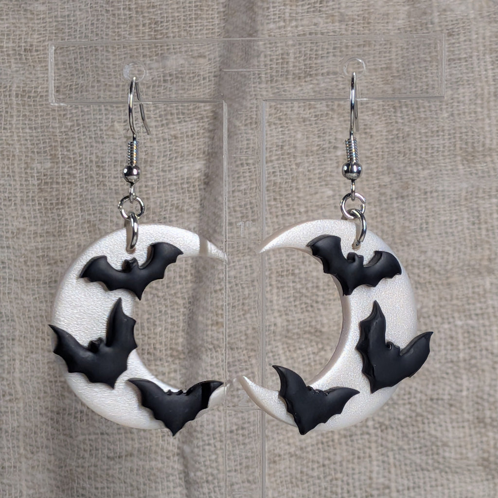 A pair of white crescent moon dangle earrings decorated with black bat silhouettes, hanging from silver fishhook wires against a textured beige fabric backdrop.
