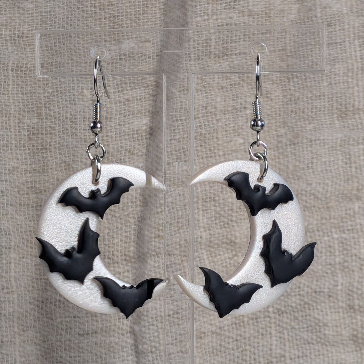 A pair of white crescent moon dangle earrings decorated with black bat silhouettes, hanging from silver fishhook wires against a textured beige fabric backdrop.