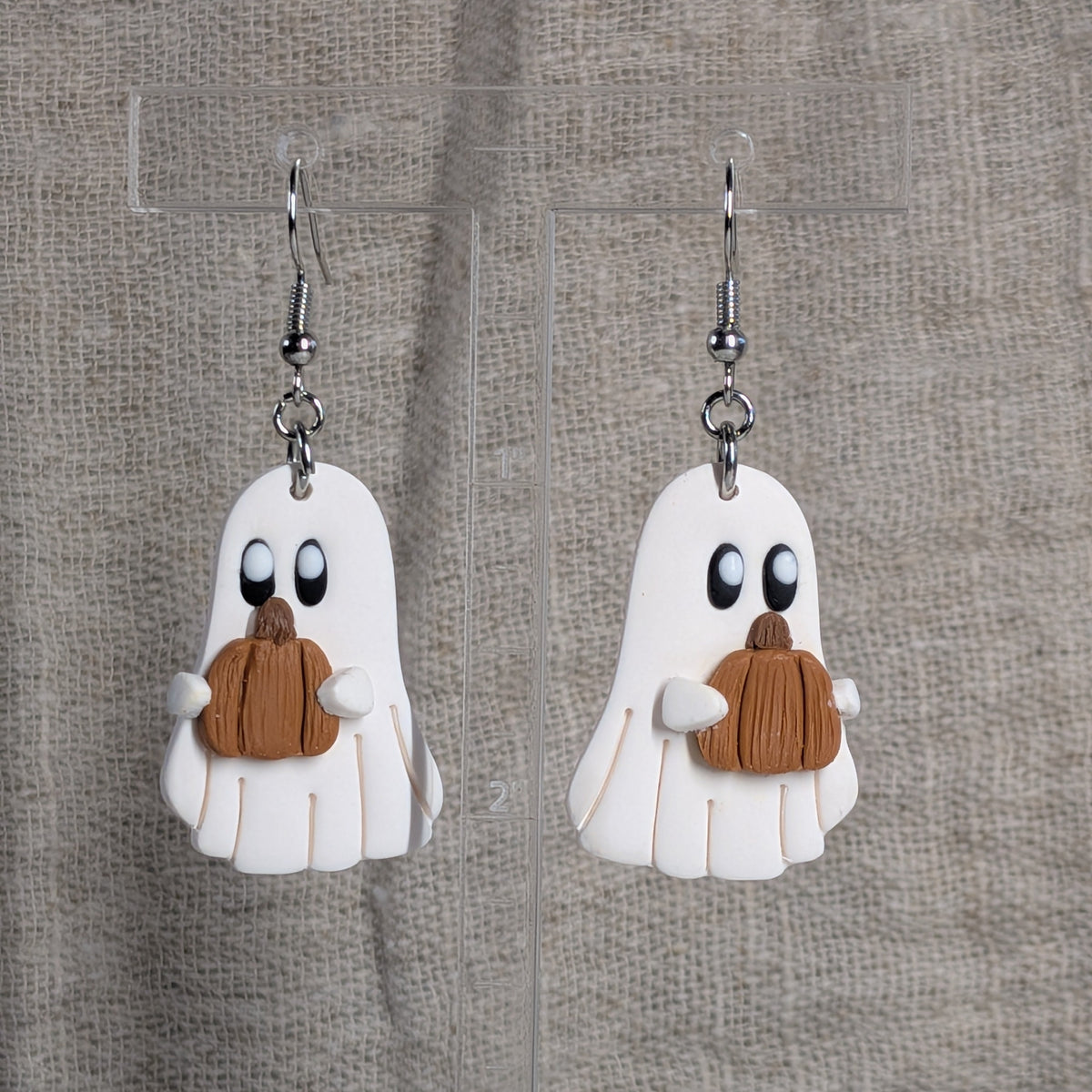 A pair of white ghost dangle earrings with brownish-orange pumpkins, displayed on a clear acrylic stand against a textured beige linen background.
