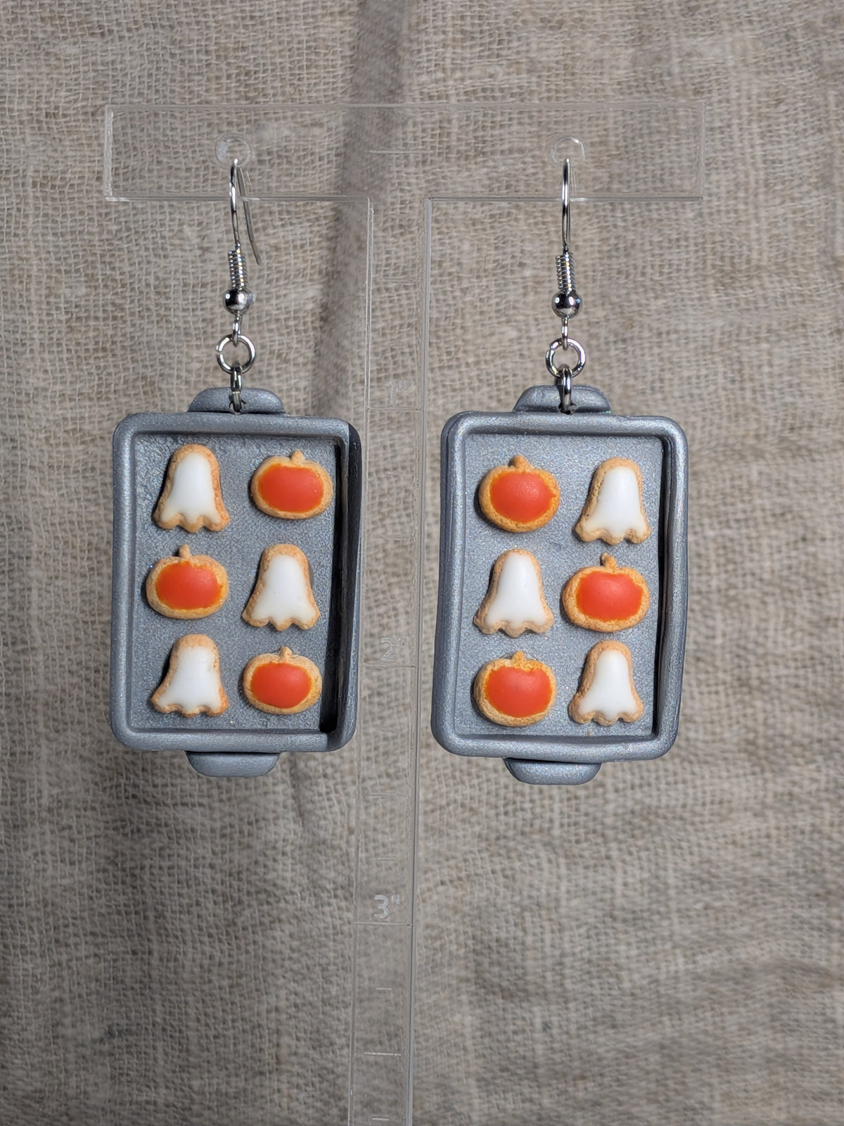 A pair of dangling earrings shaped like miniature baking trays, featuring orange pumpkin and white ghost motifs, displayed on clear acrylic stands against a textured beige fabric background.