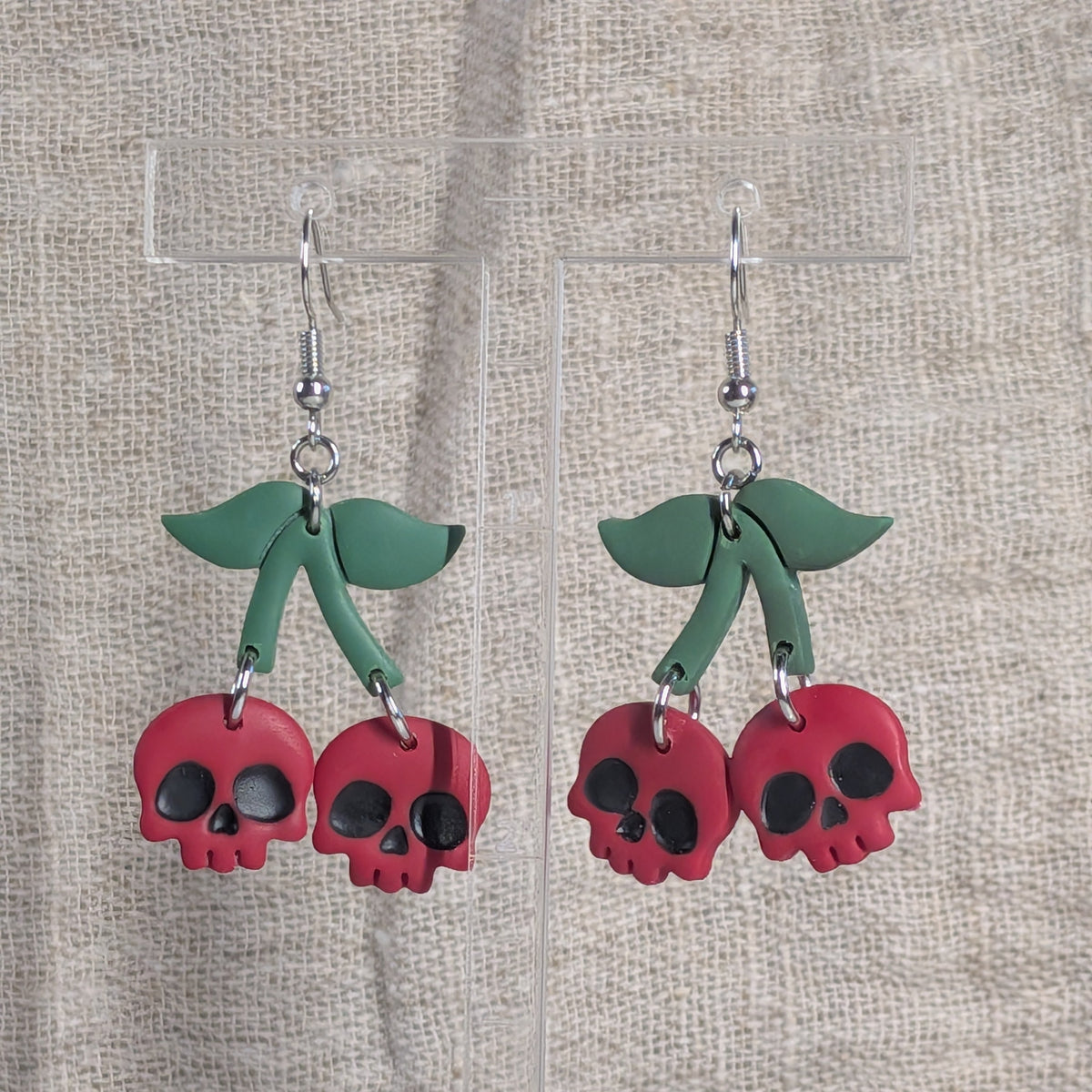 A pair of dangling earrings featuring red cherry-shaped skulls with green leaves and silver fishhook wires, displayed on clear acrylic stands against a textured beige linen background.