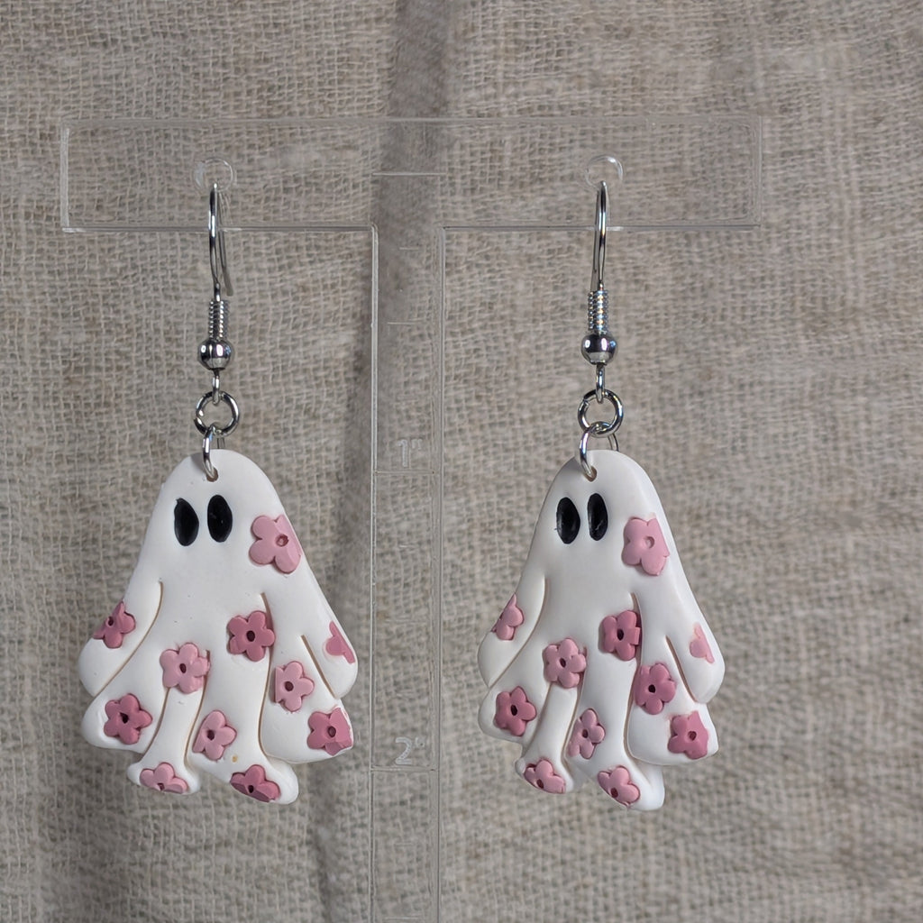 A pair of white ghost-shaped dangle earrings with pink flower details and black eyes, hanging from silver fishhook wires against a neutral textured fabric background.