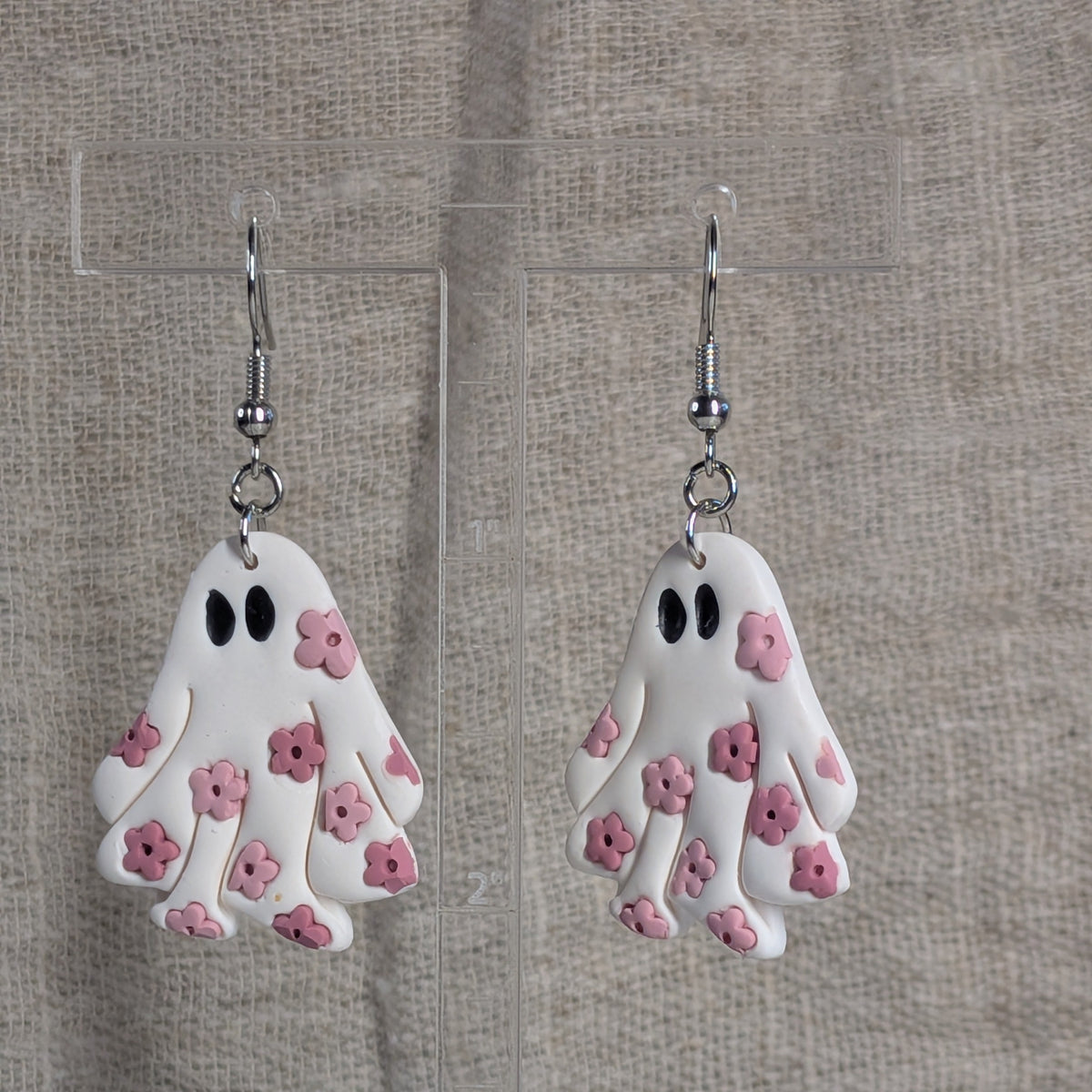 A pair of white ghost-shaped dangle earrings with pink flower details and black eyes, hanging from silver fishhook wires against a neutral textured fabric background.
