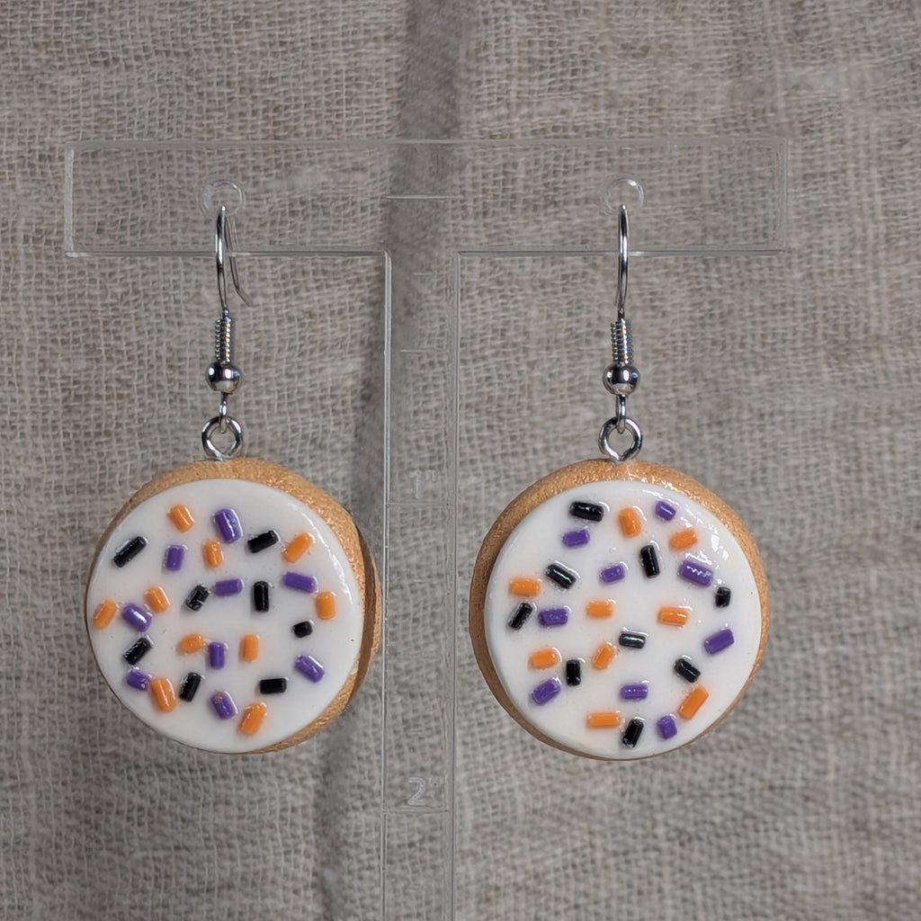 A pair of dangling earrings shaped like round frosted sugar cookies with white icing and sprinkles in orange, black, and purple, hanging from silver hooks against a textured beige fabric background.