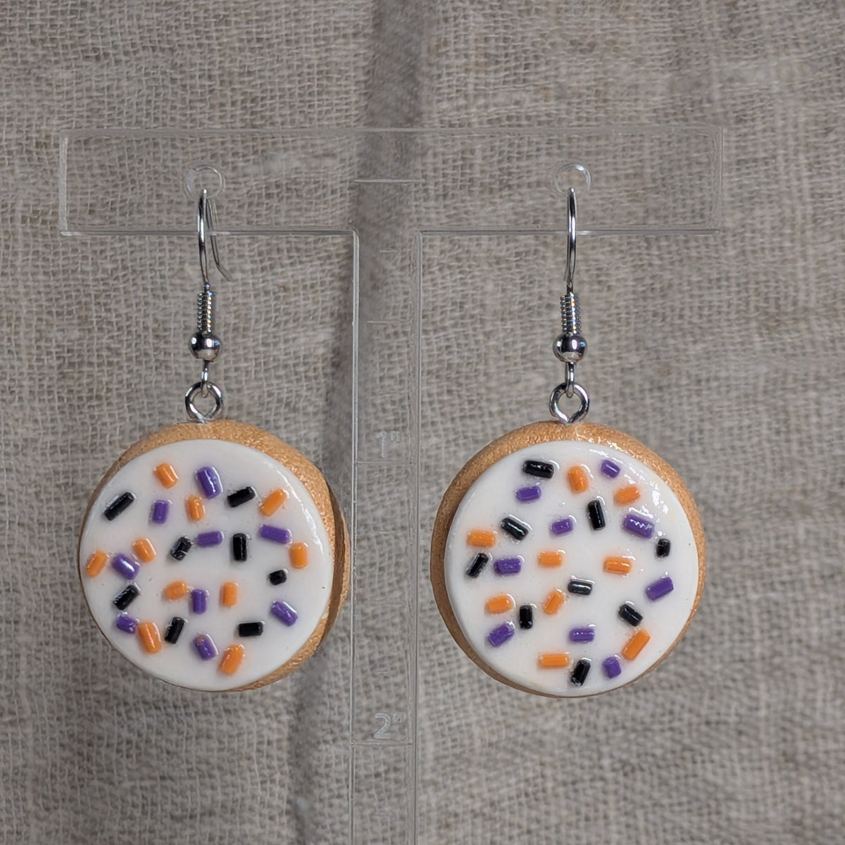 A pair of dangling earrings shaped like round frosted sugar cookies with white icing and sprinkles in orange, black, and purple, hanging from silver hooks against a textured beige fabric background.