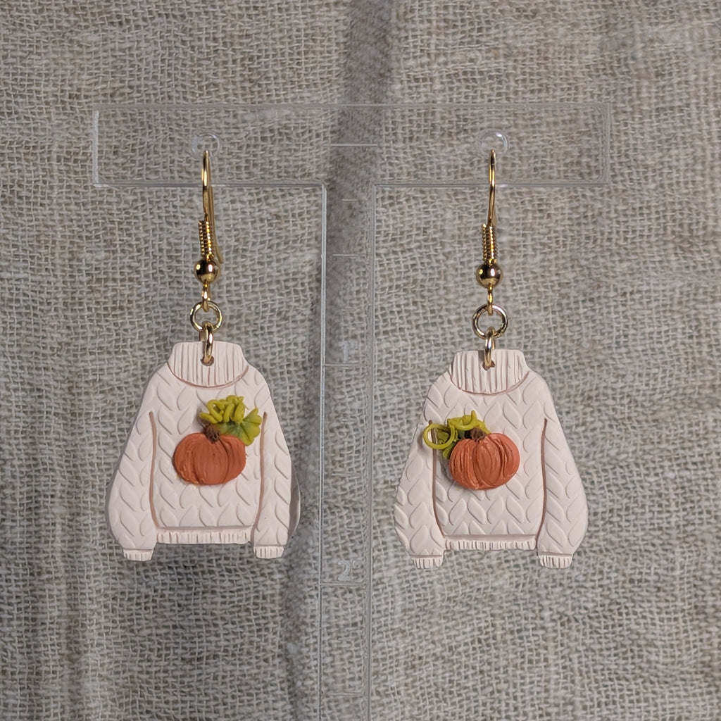 A pair of dangling earrings shaped like cream-colored cable-knit sweaters with orange pumpkin patches backed with green leaves and curly vines on the front, hanging from gold fishhook wires. They hang from a clear acrylic stand against a textured gray fabric background.