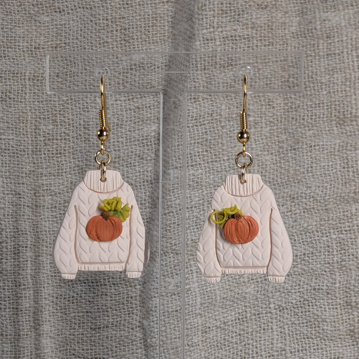 A pair of dangling earrings shaped like cream-colored cable-knit sweaters with orange pumpkin patches backed with green leaves and curly vines on the front, hanging from gold fishhook wires. They hang from a clear acrylic stand against a textured gray fabric background.