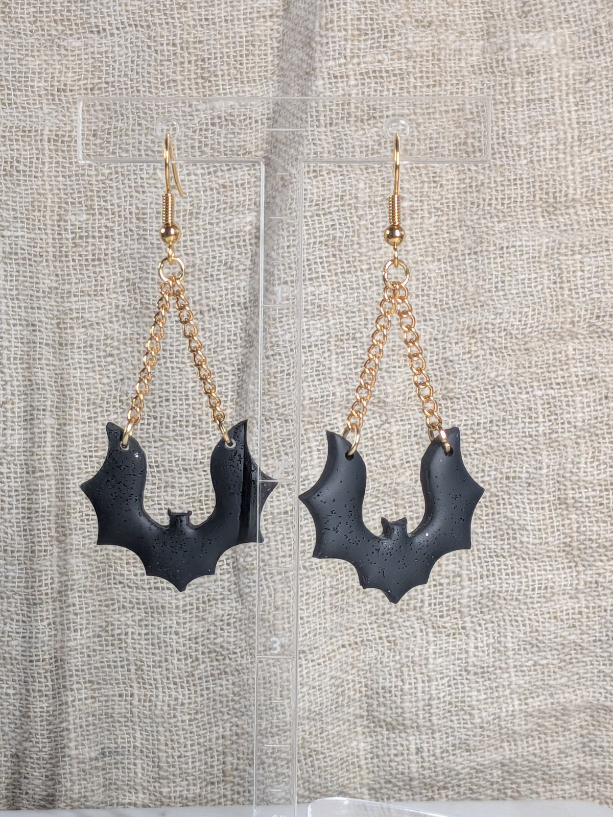 A pair of gothic-style bat drop earrings with black glitter bats hanging from gold-tone chains and fishhook wires, displayed on a clear acrylic stand against a beige linen fabric background.