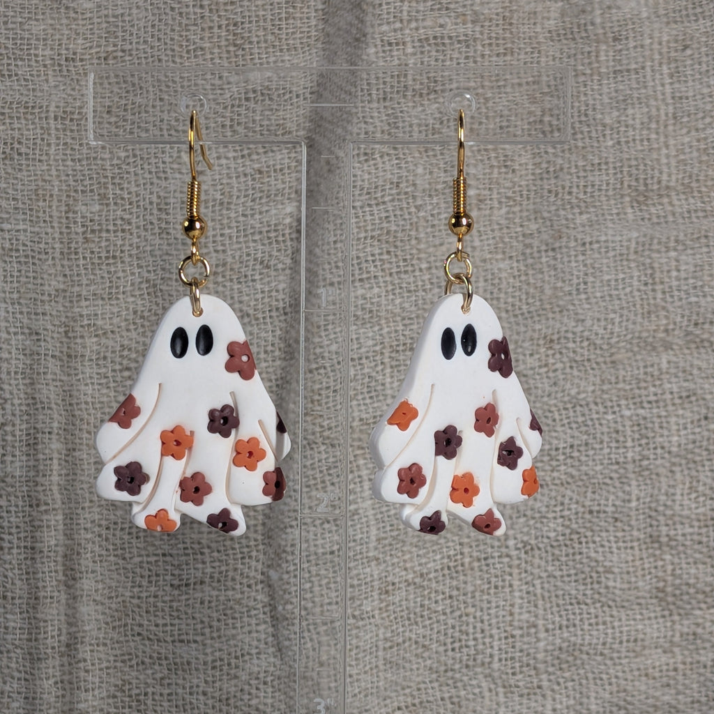 A pair of ghost-shaped dangle earrings with a white body, black eyes, and a floral pattern in orange, red-brown, and burgundy, hanging from gold-tone hooks against a textured beige fabric backdrop.