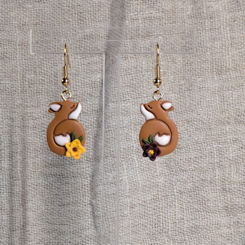 A pair of dangling earrings with terracotta colored earrings showing curled foxes with white accents and small yellow and purple flowers near the bottoms, displayed on clear acrylic stand against a textured linen background.