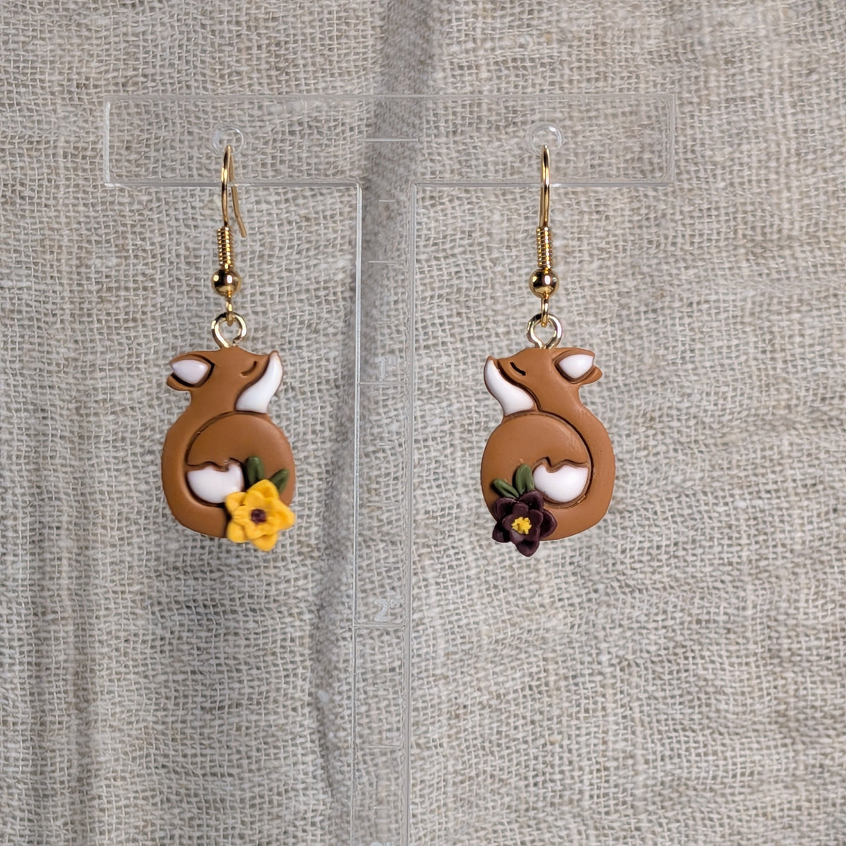 A pair of dangling earrings with terracotta colored earrings showing curled foxes with white accents and small yellow and purple flowers near the bottoms, displayed on clear acrylic stand against a textured linen background.