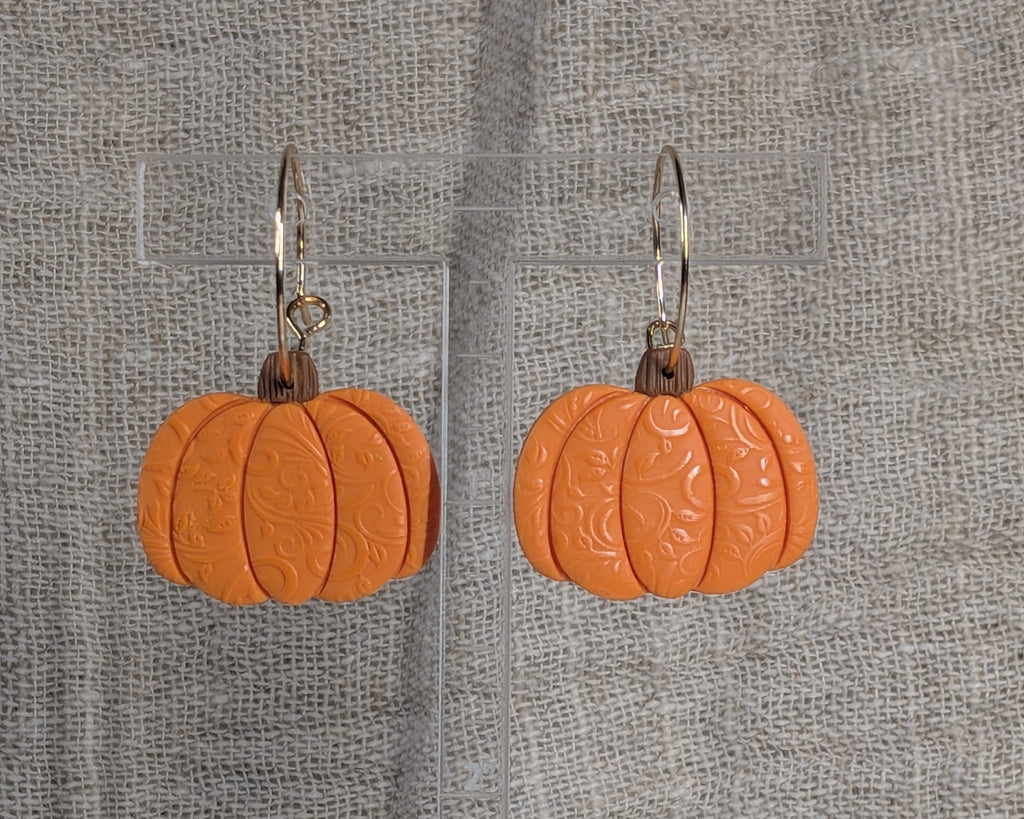 A pair of orange pumpkin-shaped dangle earrings with a subtle swirl texture on gold plated hoop wires, displayed on a clear acrylic stand against a textured gray burlap background.