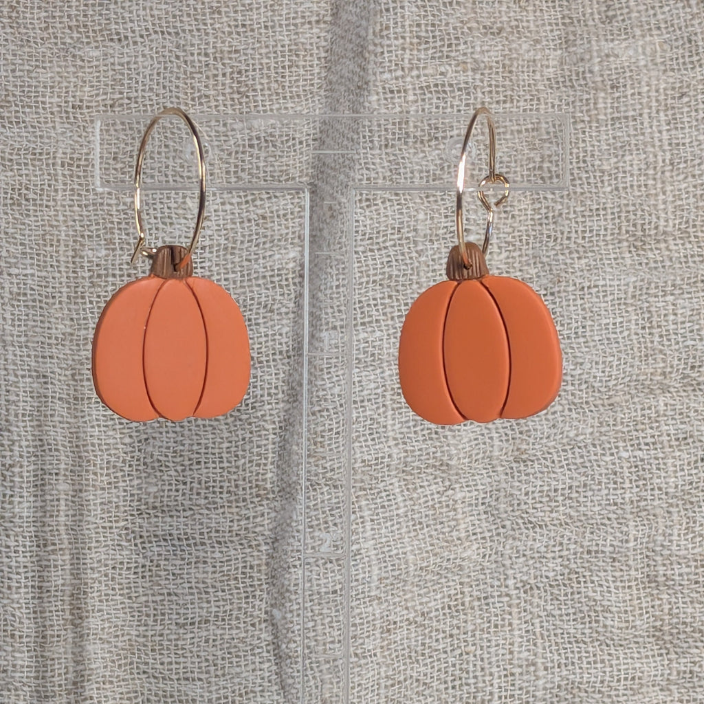 Pumpkin-shaped drop earrings with a matte terracotta orange finish and gold plated wire hooks, displayed on a clear acrylic stand against a textured beige fabric backdrop.
