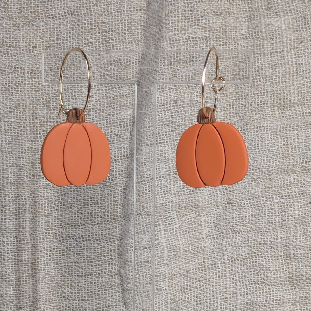 Pumpkin-shaped drop earrings with a matte terracotta orange finish and gold plated wire hooks, displayed on a clear acrylic stand against a textured beige fabric backdrop.