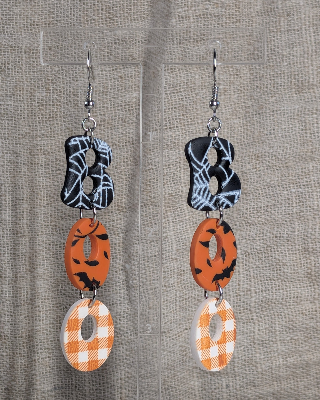 A pair of dangling earrings displayed on a clear stand against a textured beige fabric background. Each earring has a silver fishhook wire, a black and white spiderweb-patterned top disc, an orange middle disc with black bat silhouettes, and a white and orange gingham-patterned bottom disc, which together spell out the word "Boo."