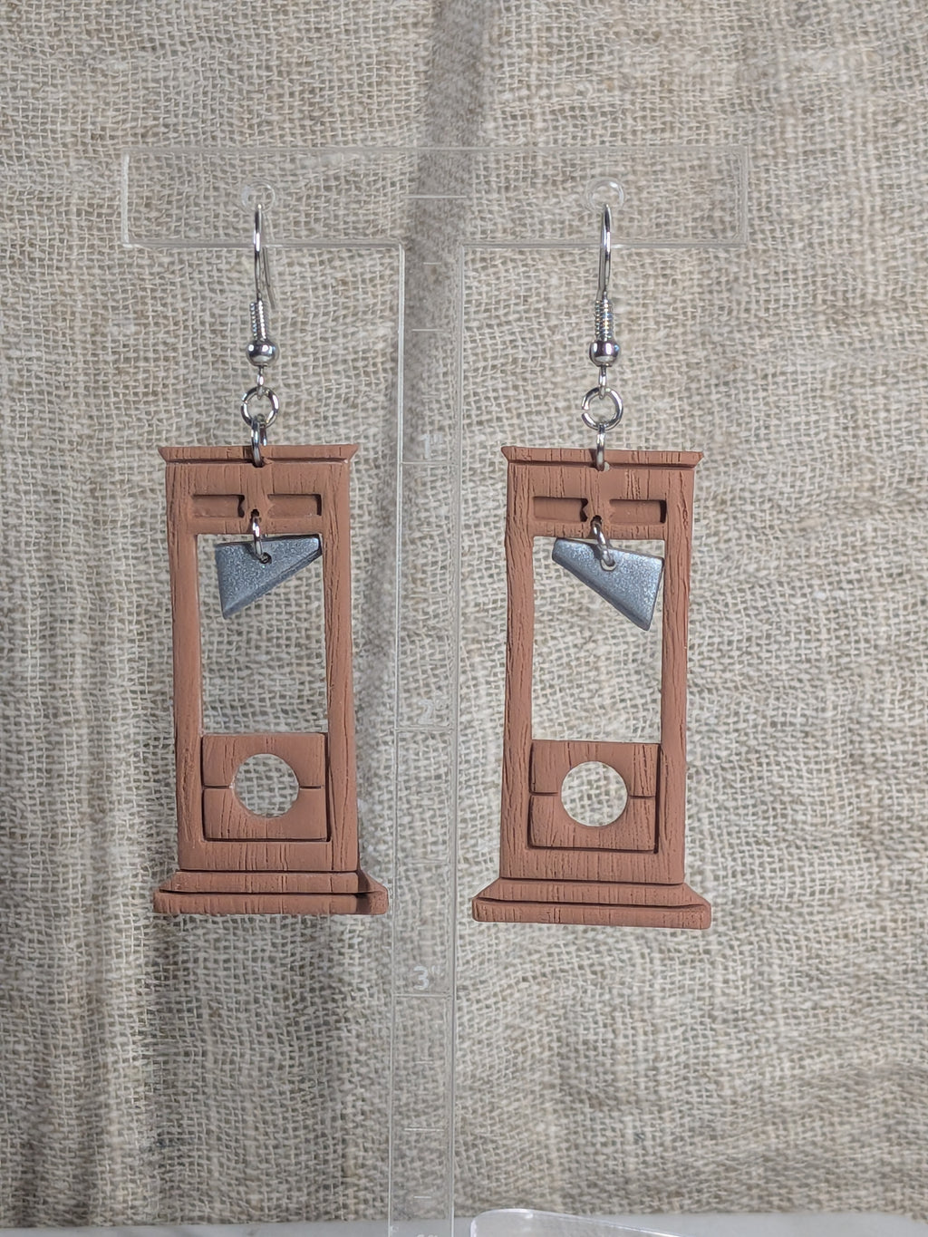 A pair of dangling earrings shaped like wooden guillotine blocks, with silver fishhook wires, displayed on clear acrylic stands against a textured beige fabric background. 