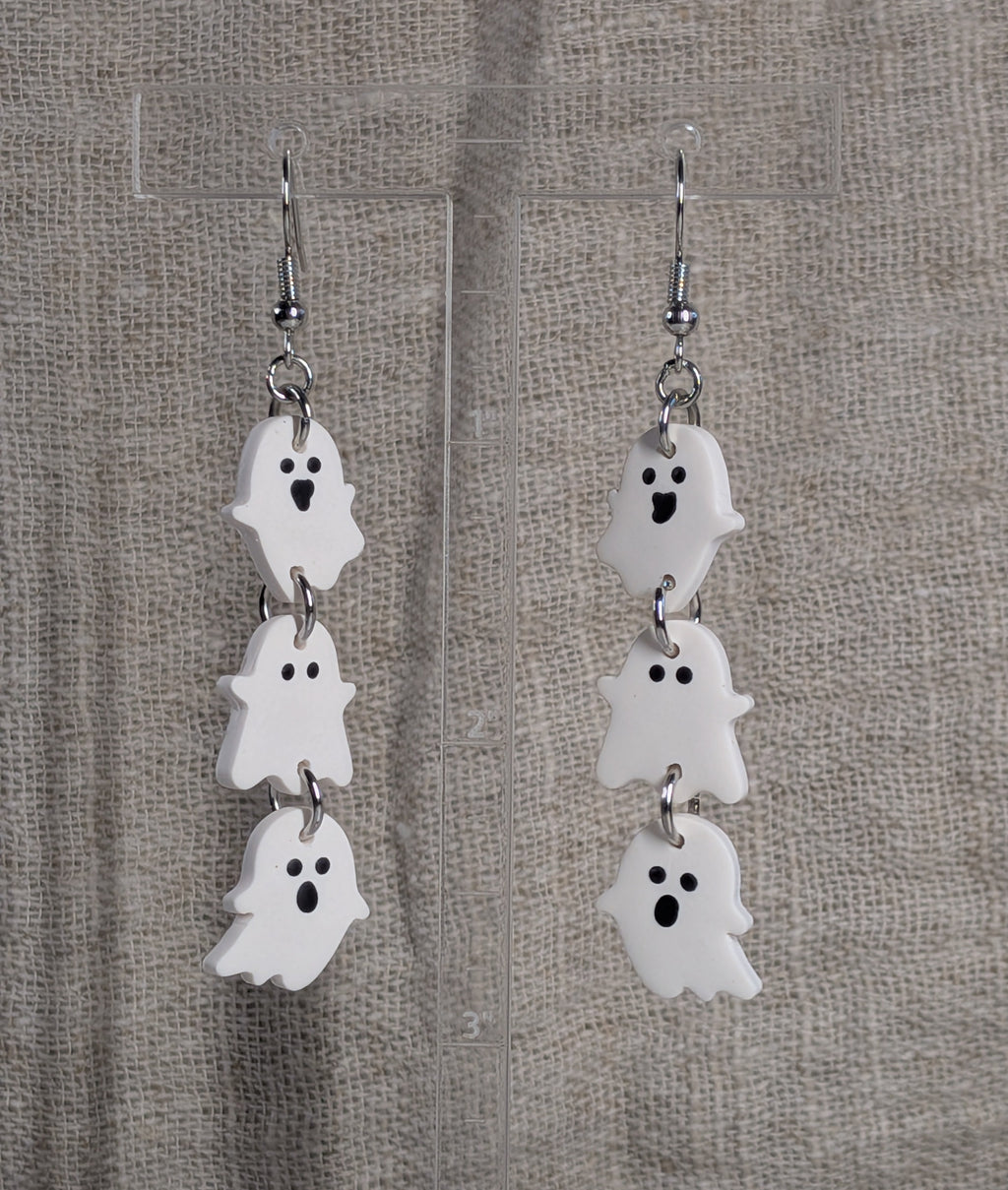 A pair of dangling ghost earrings with three stacked white ghost charms and black facial features, hanging from silver fishhook wires against a textured beige fabric background.