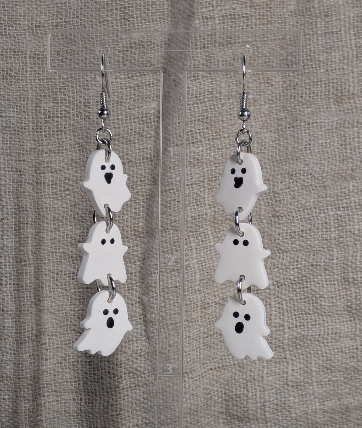 A pair of dangling ghost earrings with three stacked white ghost charms and black facial features, hanging from silver fishhook wires against a textured beige fabric background.