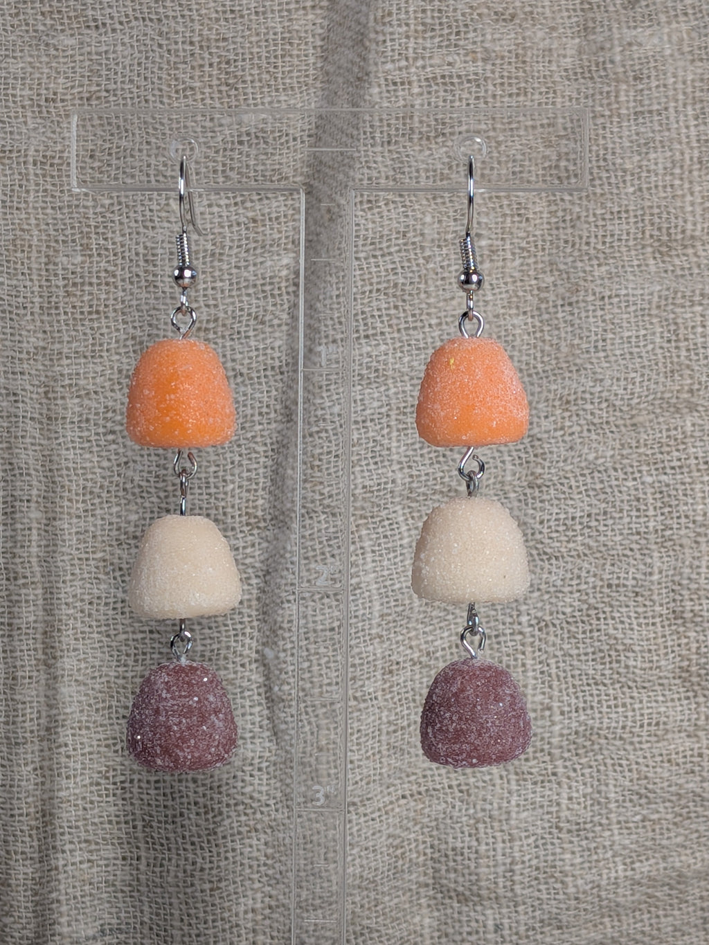 A pair of dangling earrings with three stacked gumdrop-shaped beads in orange, cream, and dusty mauve, suspended from silver fishhook wires against a textured beige fabric background.