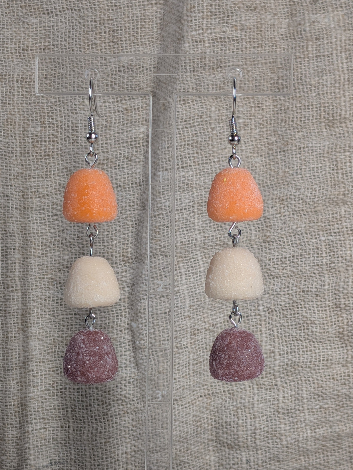 A pair of dangling earrings with three stacked gumdrop-shaped beads in orange, cream, and dusty mauve, suspended from silver fishhook wires against a textured beige fabric background.