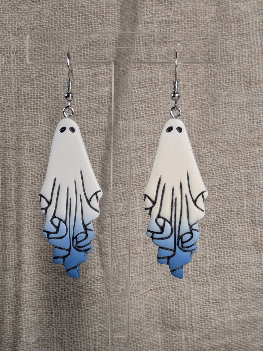 A pair of ghost-shaped dangle earrings with a white to blue gradient and black line details, displayed on a clear stand against a textured beige fabric background.