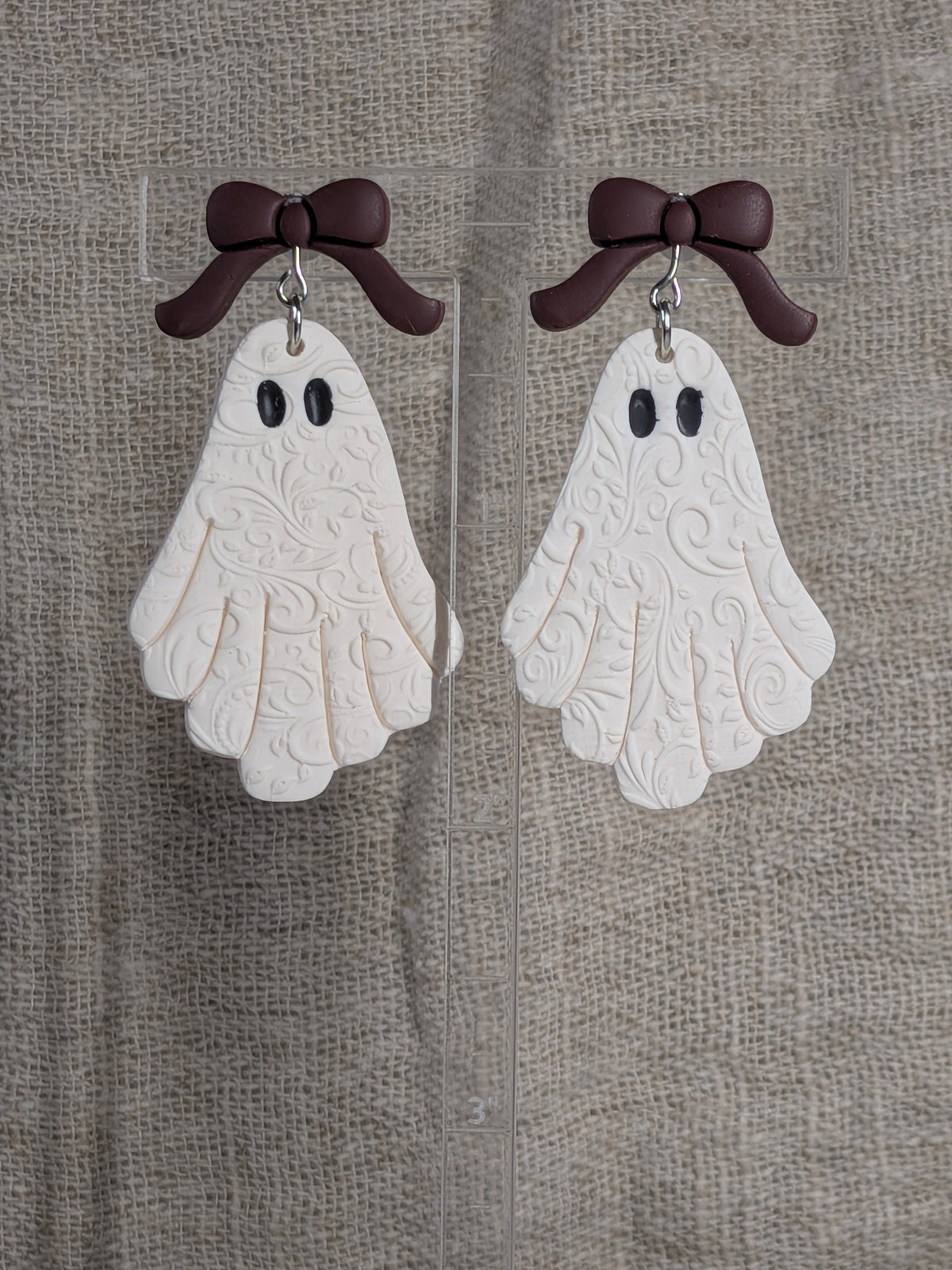 A pair of ghost-shaped earrings with embossed floral swirl patterns, featuring burgundy bows at the top and black eyes, displayed on a clear stand against a textured beige fabric background.