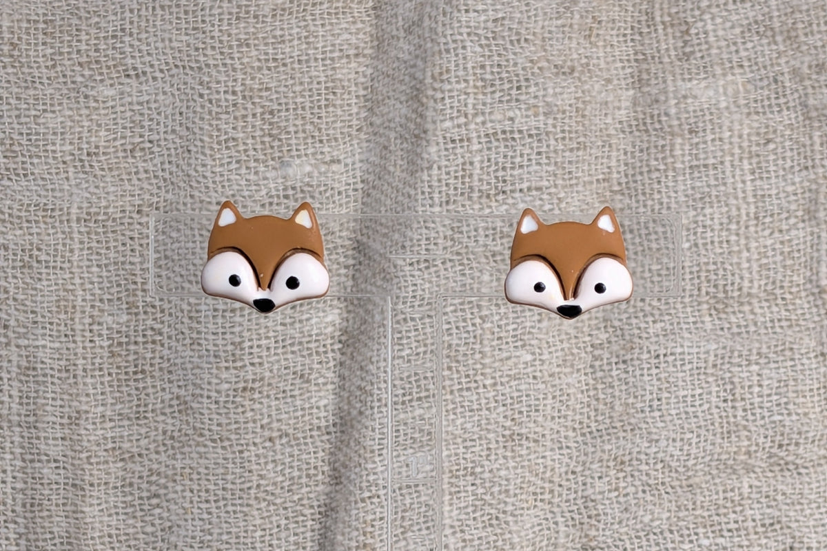 A pair of small fox face stud earrings with brown, white, and black details, displayed on a textured light gray linen background.