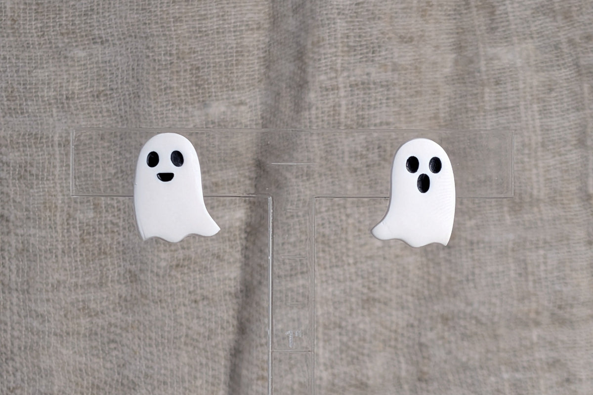 A pair of white ghost-shaped stud earrings with black eyes and mouths,  one with a smile, one with a shocked expression, displayed on a clear acrylic block against a textured beige linen background.