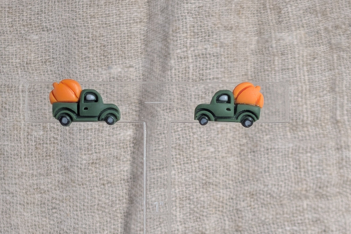 A pair of stud earrings shaped like green pickup trucks carrying orange pumpkins, displayed on a clear acrylic stand over a textured beige linen fabric background.